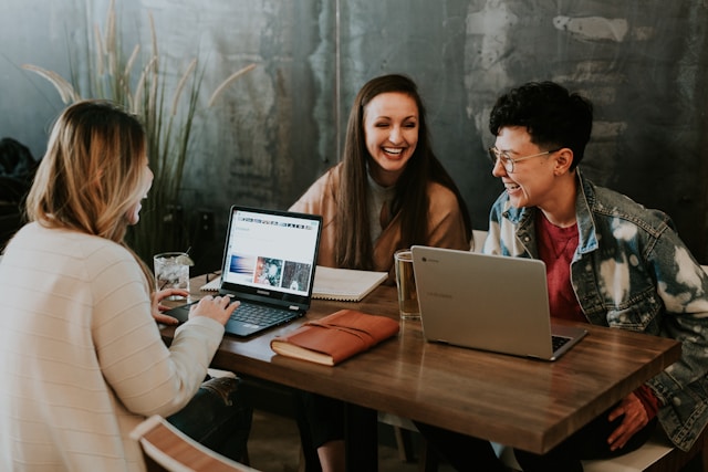 Trzy młode osoby siedzą przy drewnianym stoliku w nowoczesnej kawiarni lub przestrzeni coworkingowej. Dwie z nich korzystają z laptopów, a wszystkie są uśmiechnięte i prowadzą swobodną rozmowę. Na stole leży notes i szklanki z napojami. W tle widoczna jest ciemna, betonowa ściana z roślinami. Atmosfera jest ciepła i sprzyjająca współpracy.